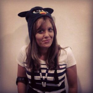 Kay Ashton stands against a plain background, smiling warmly. She wears a T‑shirt and a playful Furby‑themed hat, with her long hair falling neatly on either side of her face. The image has a filter on it. You can slightly see her crutch on her arm.
